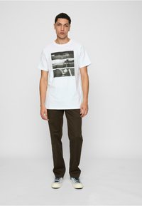 Camiseta de algodón blanca con un estampado gráfico en blanco y negro en la parte delantera, combinada con pantalones marrones y zapatillas azul claro.