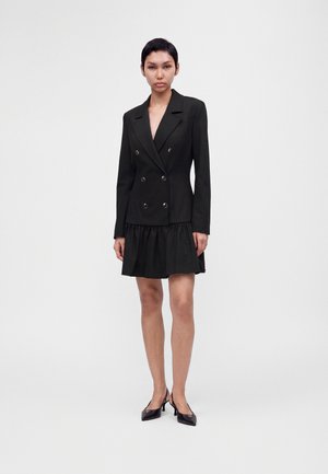 Femme aux cheveux courts et foncés portant une robe blazer noire double boutonnage avec un ourlet volanté et des escarpins noirs à bout pointu, debout sur un fond blanc.