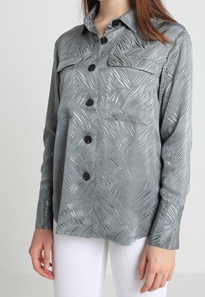 Vrouw draagt een grijs overhemd met knopen, met een subtiel abstract patroon en zwarte knopen, gecombineerd met een witte broek.