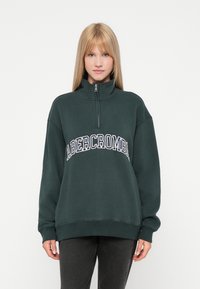 Σκούρο πράσινο φλις φούτερ με ψηλό γιακά, φερμουάρ μέχρι το ένα τέταρτο και κείμενο "ABERCROMBIE" σε λευκό και ναυτικό μπλε στο μπροστινό μέρος. Άνετη εφαρμογή.