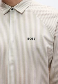 Smėlio spalvos ir baltos spalvos dryžuota marškinėliai su sagomis, su smailiu apykakliu ir juodu „BOSS“ logotipu kairėje krūtinės pusėje. Lygus audinio tekstūra.