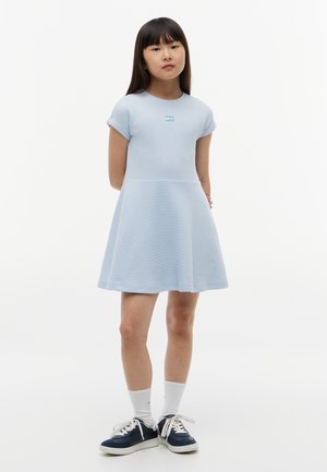 Młoda dziewczyna stoi z rękami za plecami, ubrana w jasnoniebieską sukienkę z krótkim rękawem, białe skarpetki i granatowe trampki na białym tle.