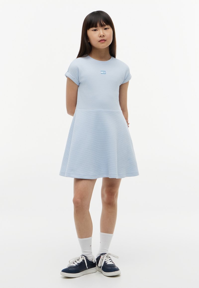 Jeune fille debout, mains derrière le dos, vêtue d'une robe bleu clair à manches courtes, de chaussettes blanches et de baskets bleu marine sur fond blanc.