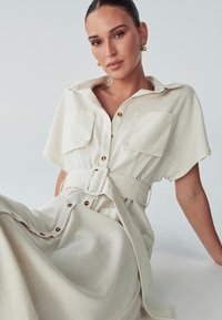 Helebeige särgidress tekstureeritud kangast, millel on lühikesed varrukad, vööga talje, kaks rinnataskut ja kuldsed nupud.