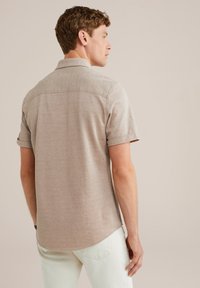 Un giovane con i capelli corti e ricci sta in piedi di lato, indossando una camicia beige a maniche corte e pantaloni bianchi su uno sfondo semplice.