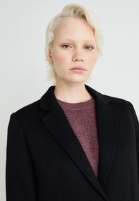 Čierny blazer s klopovým golierom, skombinovaný s textúrovaným vínovým sveterom. Tkanina vyzerá hladko s jasne definovanými švami a prispôsobeným strihom.