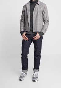 Grå, rutiga jacka över en mörk sweater, matchad med mörka jeans och grå sneakers. Uppvikt ärmslut på jeansen, lättviktsmaterial.