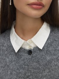 Šedý svetr s texturou, pod ním bílá košile s límečkem, s černým knoflíkem v oblasti krku.