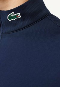 Polo bleu marine avec un col montant, un tissu élégant et un logo crocodile vert distinctif sur le col. Texture lisse.