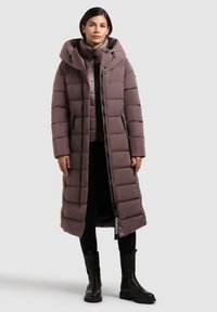 Dugi, puffer kaput u mauve boji s visokim ovratnikom i kapuljačom. Ima kvadratni uzorak, prednji zatvarač i bočne džepove. Uparen s crnim gležnjačama.