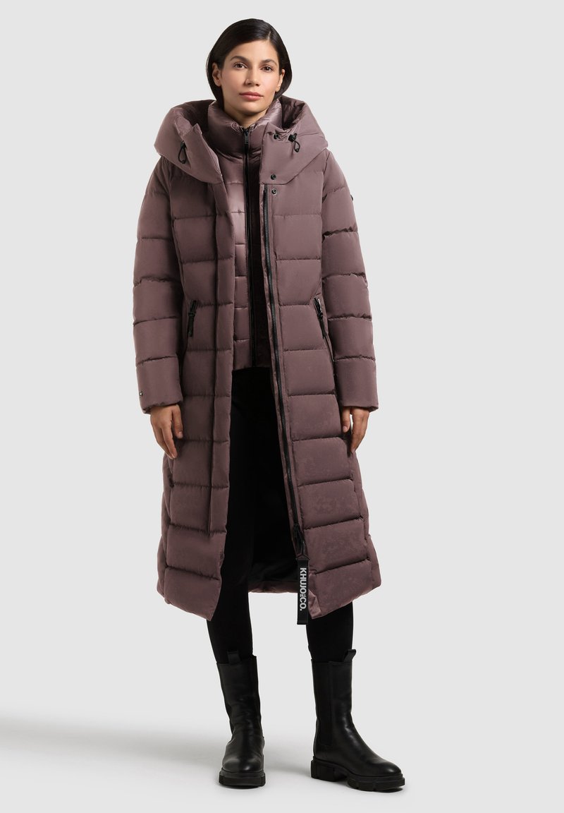 Dugi, puffer kaput u mauve boji s visokim ovratnikom i kapuljačom. Ima kvadratni uzorak, prednji zatvarač i bočne džepove. Uparen s crnim gležnjačama.