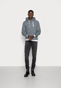 Felpa grigia con cappuccio e lacci bianchi, indossata sopra una camicia bianca, abbinata a jeans skinny neri e stivaletti neri.