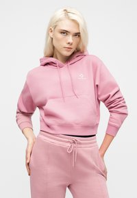 Νεαρή γυναίκα με ξανθά μαλλιά, φορώντας ροζ φούτερ Converse και αντίστοιχο παντελόνι φόρμας, με τα χέρια στη μέση, κοιτάζει προς την κάμερα.