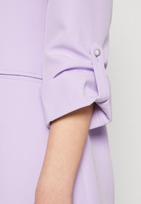 Manches en tissu lavande avec revers roulés, ornées d'un détail bouton décoratif. Texture lisse et design ajusté pour une apparence élégante.