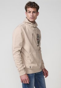 Felpa beige con collo a imbuto, caratterizzata da una grafica di teschio sul petto. Realizzata in un materiale morbido con tasca a marsupio.