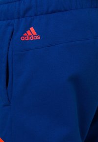 Modré sportovní tepláky s hladkou texturou, které mají oranžové logo Adidas a boční kapsy pro praktičnost.