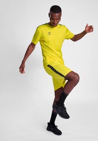 Helles gelbes Sportshirt und Shorts mit schwarzen Seitenakzenten, getragen mit schwarzen Turnschuhen und Socken; leichtes, atmungsaktives Material.