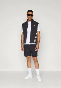 Muškarac u crnom bezrukavom prsluku, bijeloj majici, crnim šortsevima, bijelim čarapama, bijelim tenisicama i tamnim naočalama, na jednostavnoj pozadini.