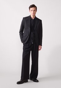 Zwart pak met een getailleerde blazer en wijde broek, beide in een gladde stof. De blazer heeft een enkele knoopsluiting en een notch revers.