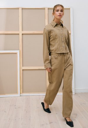 Femme marchant en intérieur portant une veste beige à clous et un pantalon large assorti avec des chaussures noires à bout pointu devant un fond de cadre en bois.