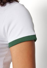 Valkoinen lyhythihainen t-paita, jossa vihreä resorireuna. Kangas näyttää sileältä, istuva malli korostaa olkapäiden ja käsivarsien muotoa.