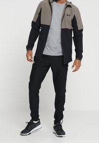 Herren-Sportoutfit: schwarze und graue Zip-Jacke mit farblich abgesetzten Schultern und Brustbereich, graues T-Shirt, schwarze Hose mit weißem Seitenstreifen, schwarze Turnschuhe.