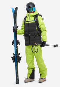 Skiausrüstung bestehend aus einer schwarzen und neongrünen Steppjacke, passenden Hosen, schwarzem Helm, verspiegelten Skibrillen, wobei blaue Skier und Stöcke gehalten werden.