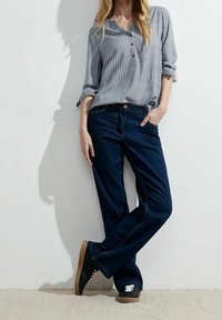 Gestreept grijs langarmshirt met knopen, donkerblauwe spijkerbroek en zwarte sneakers met witte veters. Model leunt tegen een muur.