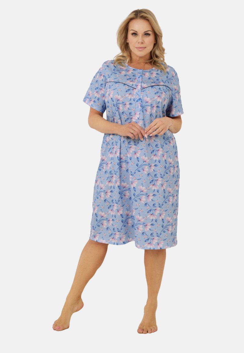 Kobieta stojąca boso w niebieskiej, kwiecistej sukience do kolan z krótkimi rękawami i zapięciem na guziki, z dłońmi złożonymi na piersi.