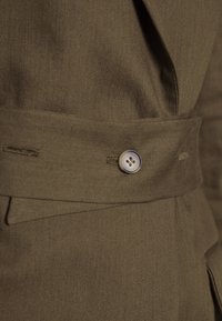 Gros plan sur une ceinture en tissu marron avec un système de fermeture à un seul bouton sur un vêtement, mettant en valeur la texture et les détails de couture.