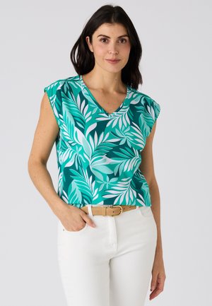 Vrouw draagt een mouwloze groene en blauwgroene top met bladmotief, witte broek en een beige riem, staand met één hand in de zak.