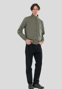 Olivgrön lättviktsjacka med ståkrage, dragkedja och två sidofickor, i kombination med svarta jeans och grå skor.