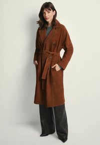 Brązowy zamszowy płaszcz trench z podwójnymi guzikami, paskiem w talii i dwoma bocznymi kieszeniami. Modelka nosi ciemne spodnie i dzianinowy top.