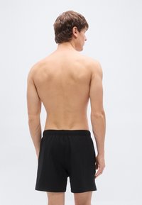 Μαύρα σορτς με ελαστική μέση, απλή υφή, μήκος μέχρι το μέσο του μηρού, από πίσω σε φόντο ανοιχτού γκρι.