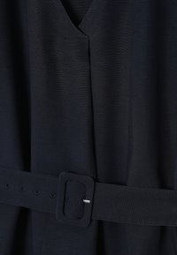 Tissu bleu marine foncé avec une ceinture assortie comportant une boucle rectangulaire et plusieurs œillets, présenté en gros plan sur un vêtement.