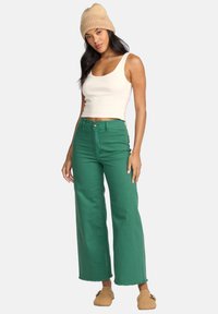 Top in crema, jeans verdi a gamba larga e un berretto a coste beige. I jeans hanno un taglio alto in vita e orlo grezzo; le sneakers completano il look.