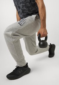 Grå træningsbukser med logo, holder en sort kettlebell, iført sorte sportssko, mørk skjorte med camouflage-mønster, i en lungestilling.