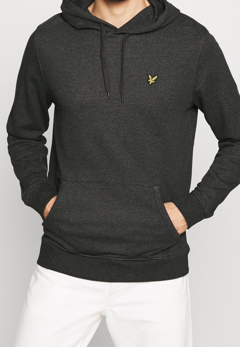 Homme portant un sweat à capuche gris foncé avec un petit logo oiseau jaune sur la poitrine et les mains dans la poche avant, associé à un pantalon blanc.