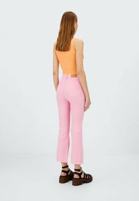 Rosa, ausgestellte Jeans mit rohem Saum, kombiniert mit einem orangefarbenen gerippten Tanktop. Das Modell trägt schwarze, geschnürte Sandalen. Rückansicht.