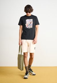 Black graphic t-shirt with a colorful urban print, cream shorts with a side pocket, green tote bag with text, and multicolored sneakers.