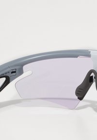 Sportsonnenbrille mit einem grauen Kunststoffrahmen, lila Gläsern und schwarzen Gummibeschlägen. Sie zeichnet sich durch ein elegantes Design mit geschwungenen Bügeln für Komfort aus.