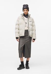 Puffy beige jas met zwarte accenten, grijze leren midi rok met een zijsplit, zwarte beanie en sneakers, wat een gelaagde look creëert.