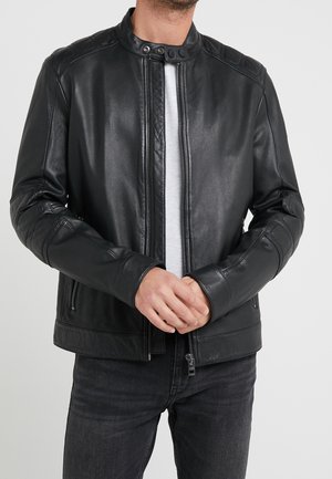 Mann trägt eine schwarze Lederjacke mit Reißverschlusstaschen über einem weißen Hemd und schwarzen Jeans, die Hände sind vor der Taille verschränkt.