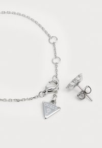 Zilveren kettingarmband met driehoekige "GUESS?" bedel en karabijnhaak, vergezeld van een bijpassende zilveren studs. 