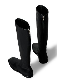 Botas de cuero negro hasta la rodilla con suelas planas, cremalleras laterales y textura suave. Presentan una punta redondeada y detalles mínimos de costura.