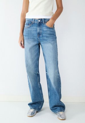 Person, die lockere hellblaue Jeans, ein weißes ärmelloses Oberteil und silberne Turnschuhe trägt, steht an einer weißen Wand.