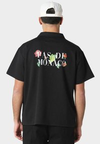 Svart kortermet skjorte med blomsterdesign og teksten "PAS DE MONACO" i hvitt. Avslappet design, laget av myk stoff.