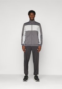 Mand står iført en grå og lysegrå Puma træningsdragt med lynlåsejakke og sorte sneakers mod en ensartet lys baggrund.