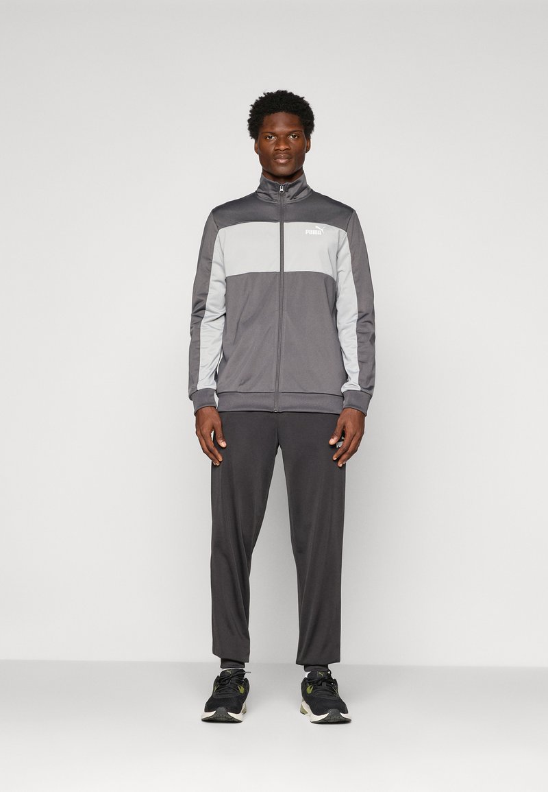 Mand står iført en grå og lysegrå Puma træningsdragt med lynlåsejakke og sorte sneakers mod en ensartet lys baggrund.