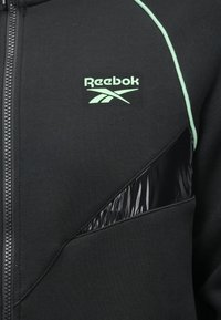 Melna zip-up jaka ar zaļo Reebok logotipu, akcentēta ar spīdīgām melnām paneļiem un kontrastējošiem zaļiem šuvju diegiem.